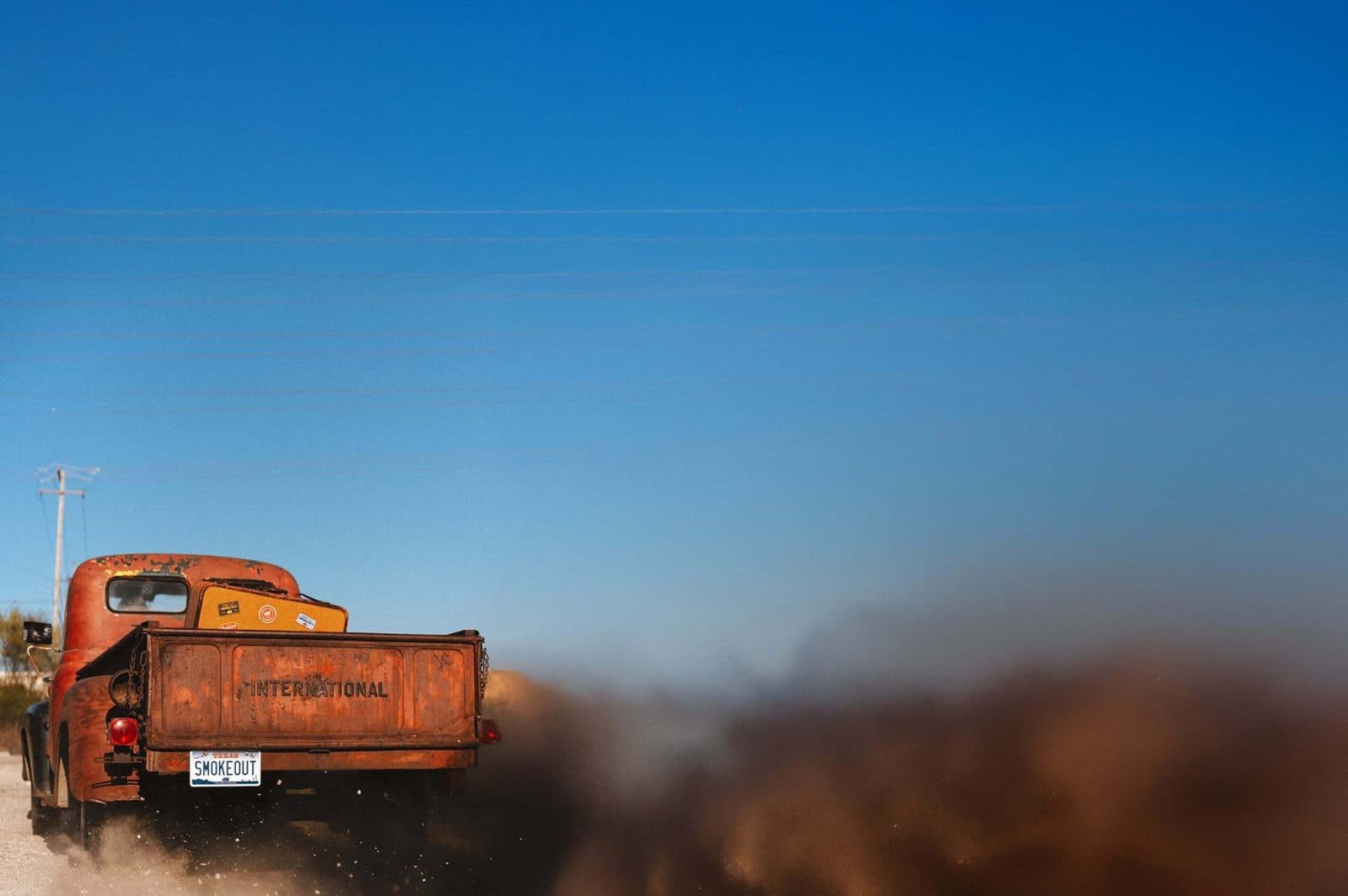 Rear view of a rusty, antique pickup truck driving away down a dirt road with a license plate that says smoke out