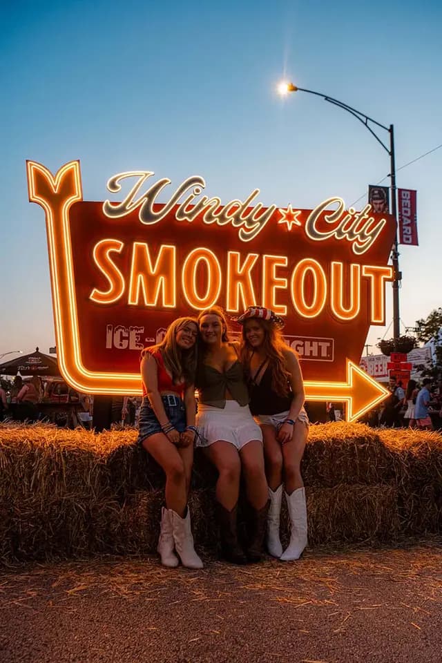 3 girls in cowboy boots sitting on a hay stack in front of a neon Windy City smokeout sign